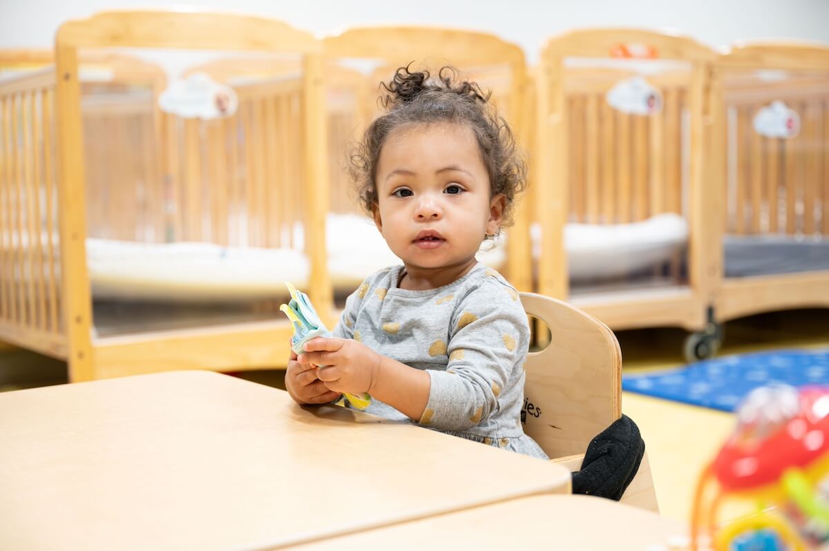 12 Young child sitting at a table in a nursery classroom, holding a soft fabric toy with cribs in the background.