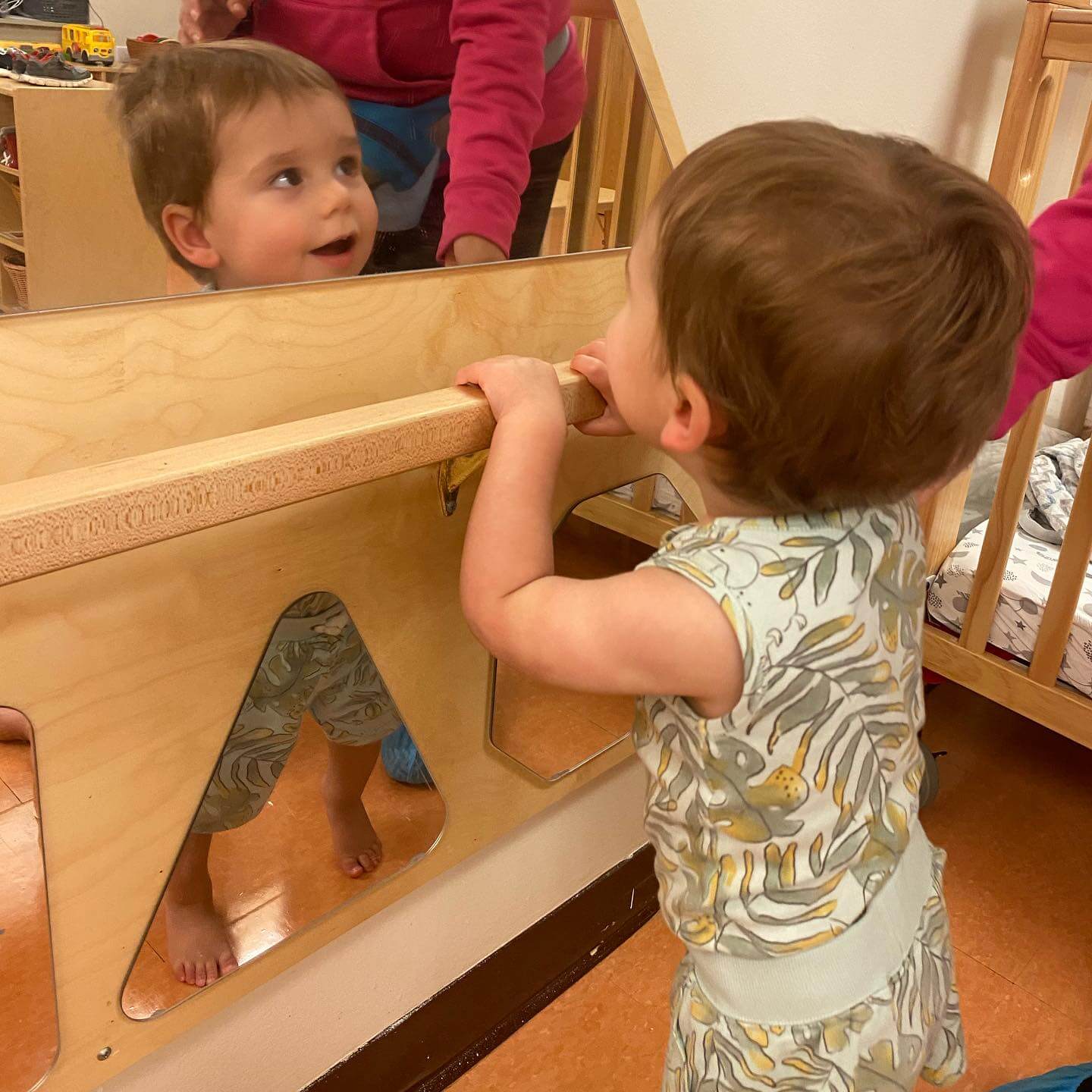 374204270_843527450908326_2212029364450493503_n Young child standing at a wooden mirror station, looking at their reflection and holding the rail.