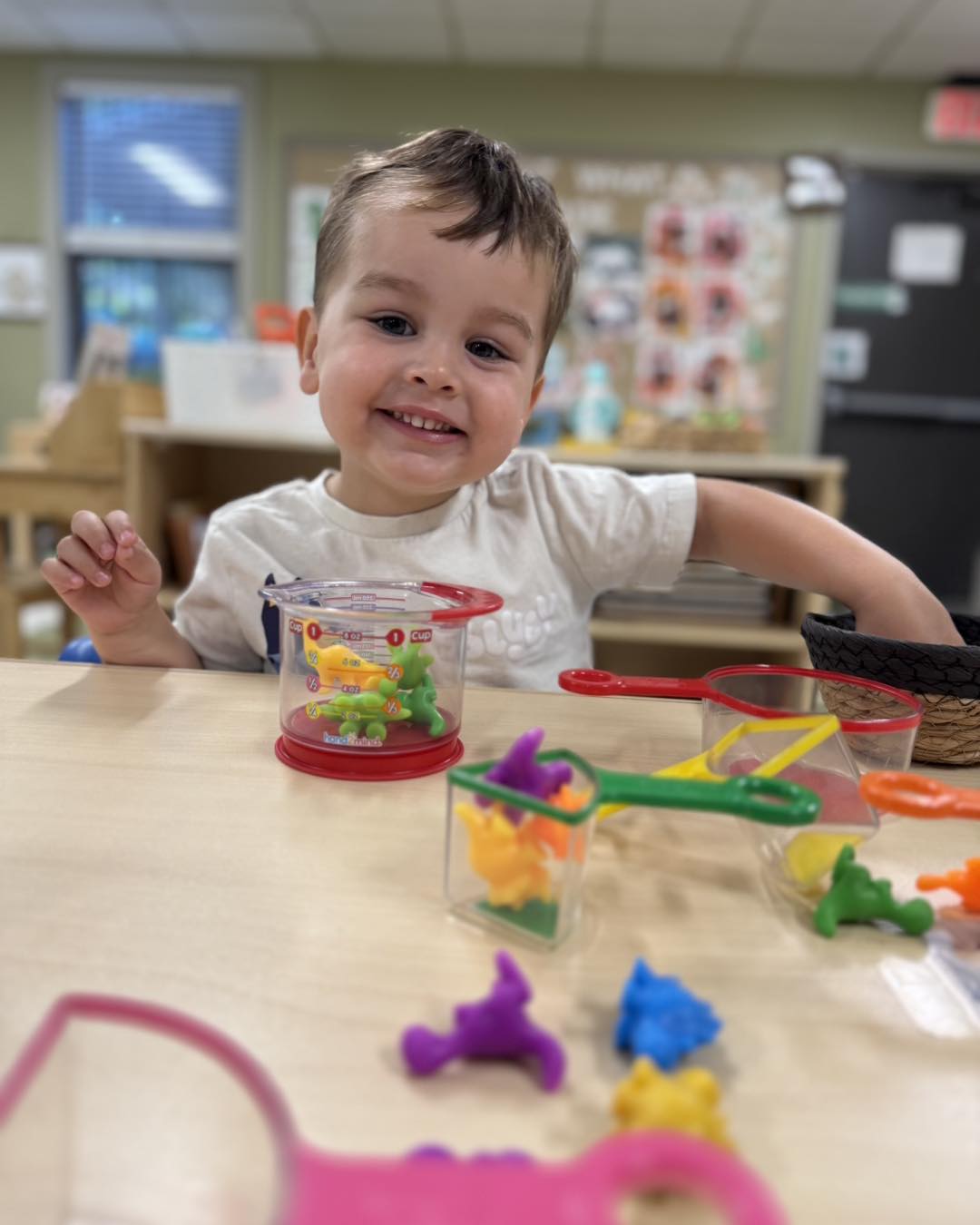 Infant_Plan_4 Smiling young child at a classroom table playing with colorful counting bears and measuring cups during a hands-on learning activity