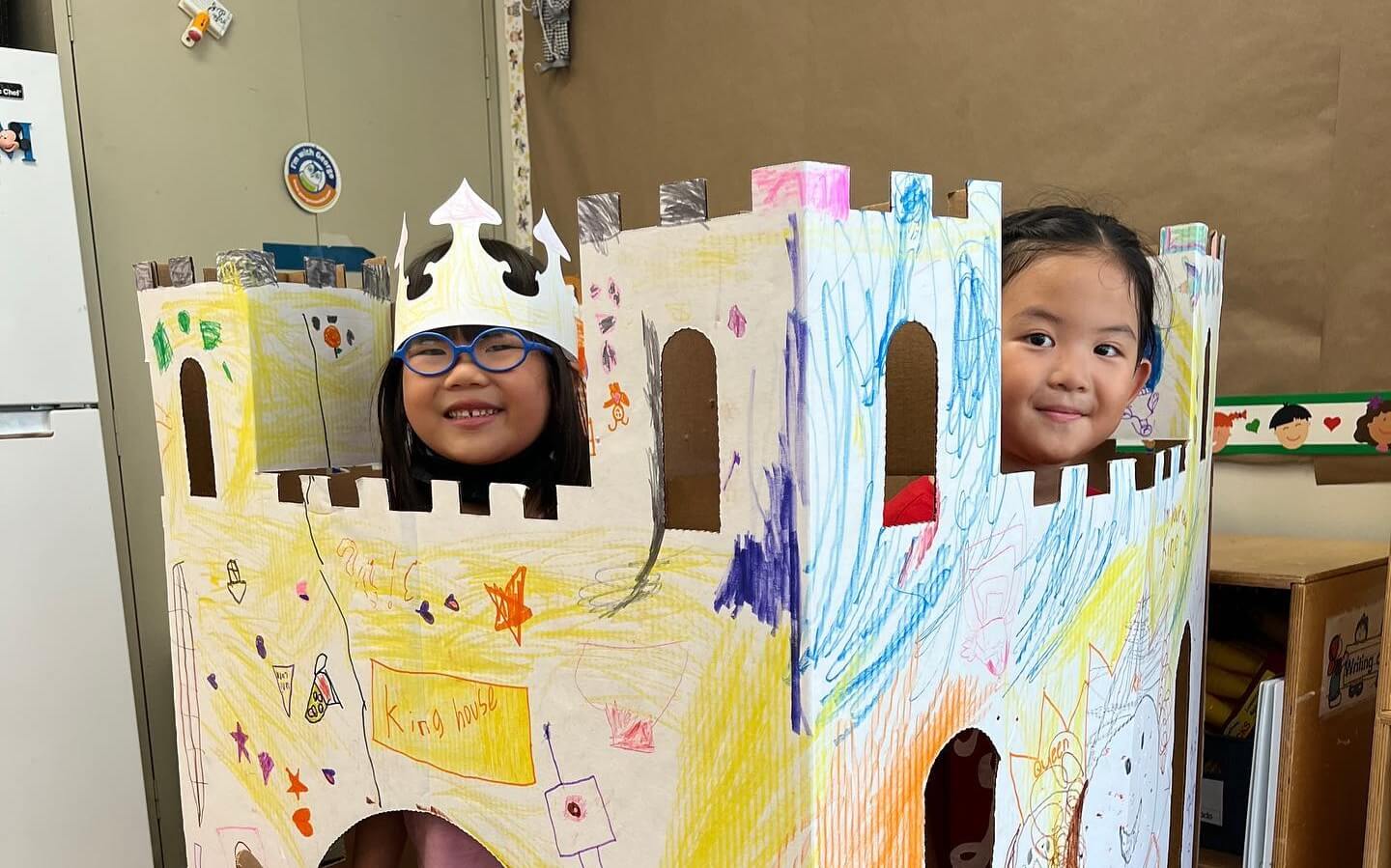 Two children playing inside a large cardboard castle decorated with colorful drawings, one wearing a paper crown.