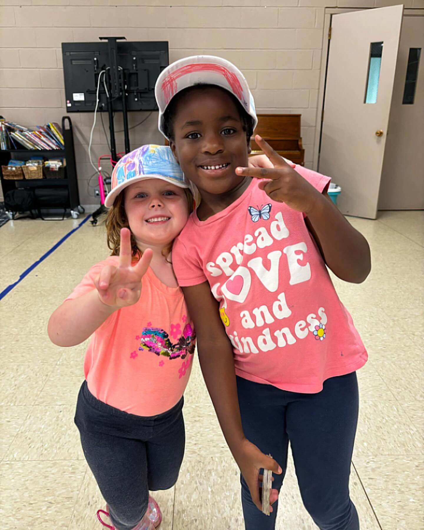 Two young girls smiling and making peace signs indoors, both wearing colorful hats and pink shirts.