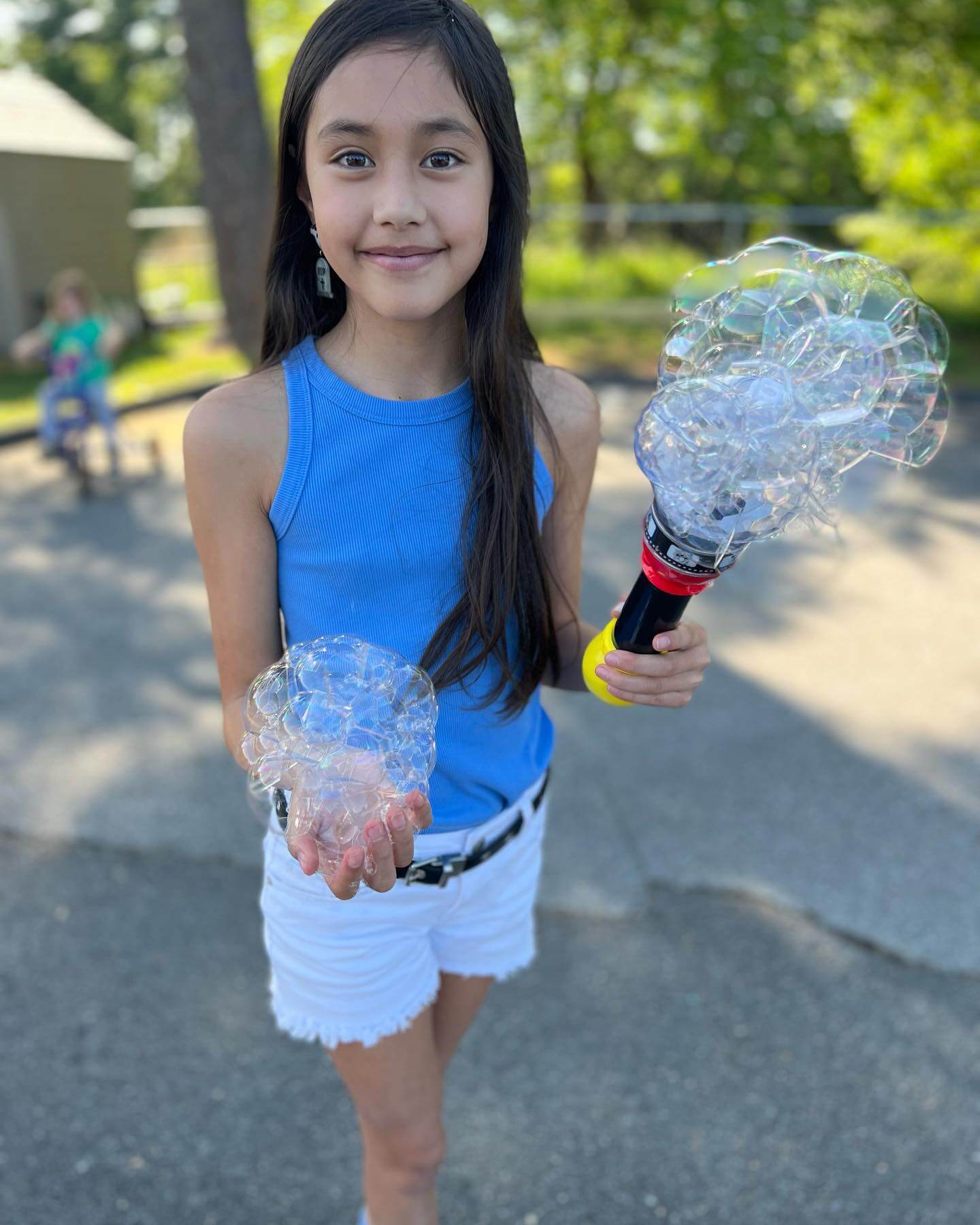 Before_5 Girl holding a bubble wand outdoors, wearing a blue sleeveless shirt and white shorts, showing off bubbles.