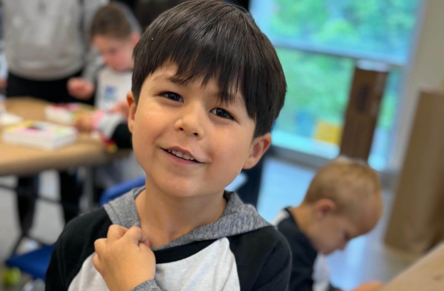 Before_6 Boy smiling indoors in a classroom setting with other children working in the background