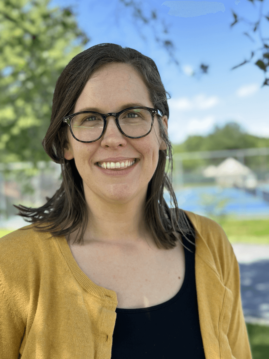 Director Emily Carmody A woman with glasses and a mustard yellow cardigan smiling at the camera, standing outside with blurred trees in the background.