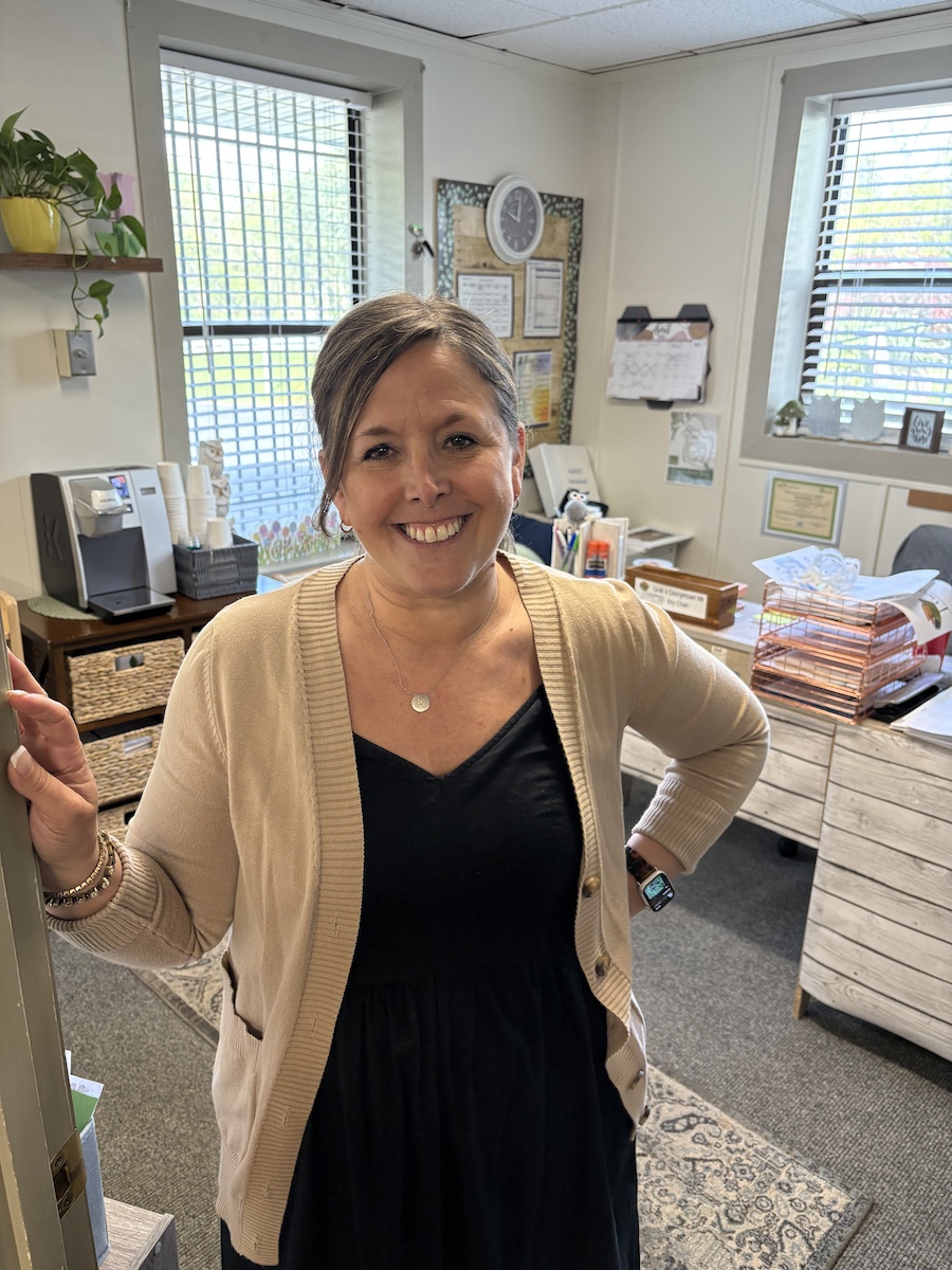 Director Melanie Morillo Woman standing in an office, smiling warmly, wearing a beige cardigan over a black dress.
