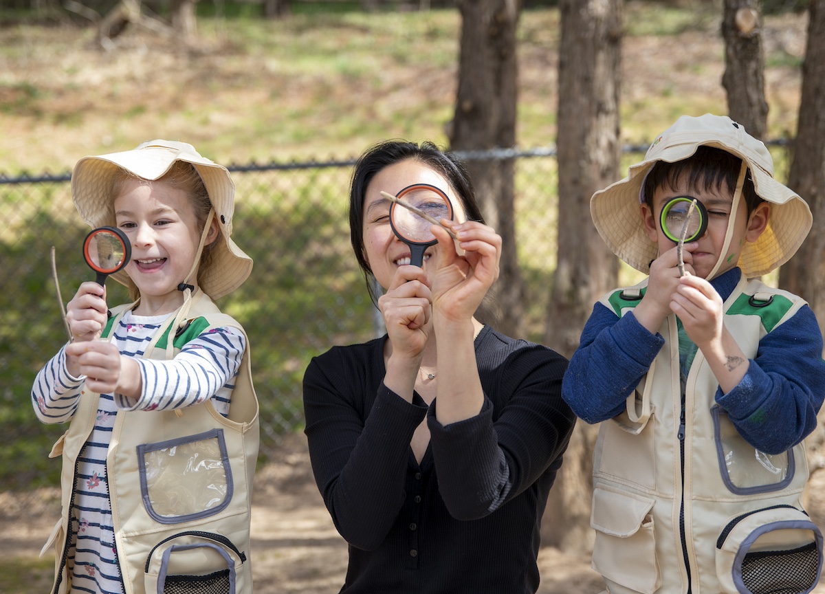 Two children and a teacher holding magnifying glasses, looking through them outdoors while smiling.