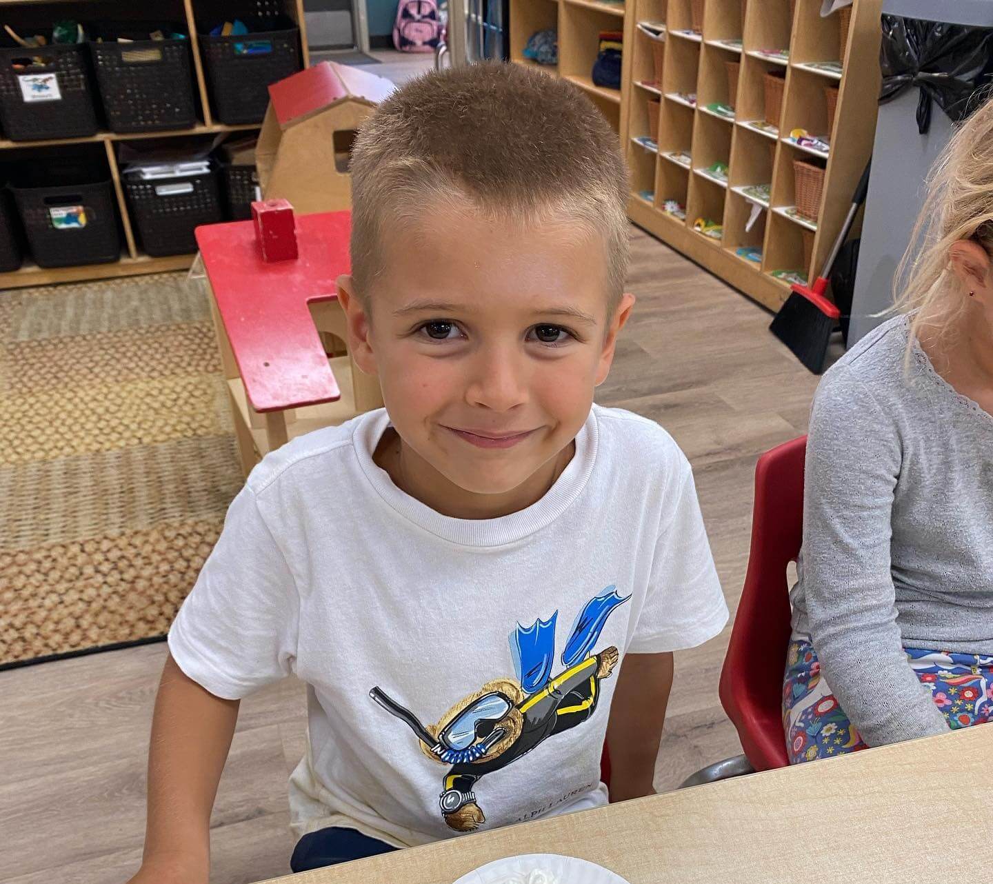 First_1 Young boy with short hair wearing a white t-shirt with a colorful scuba diver graphic, smiling while sitting at a table