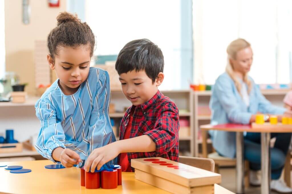 First_3 Two children working together with Montessori-style educational toys on a classroom table, one in a blue-striped shirt and the other in a red plaid shirt.