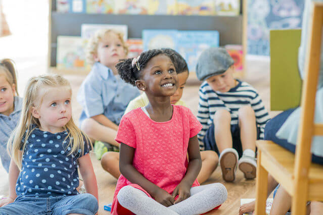 First_4 Group of young children sitting cross-legged on a classroom floor, smiling and looking toward a teacher, with one girl in a bright pink shirt in the center