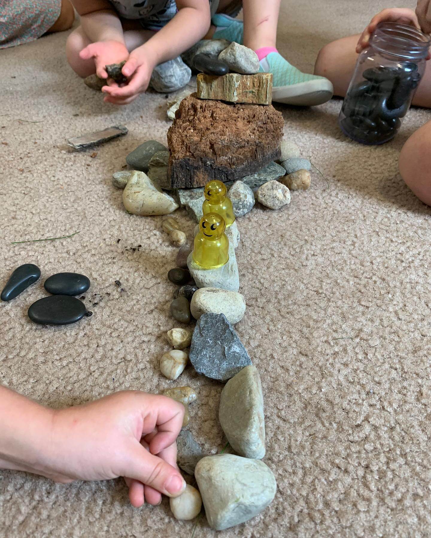 Kindergarten_1 Children sitting on the floor arranging rocks and natural materials into a path with small yellow figurines placed in the center.