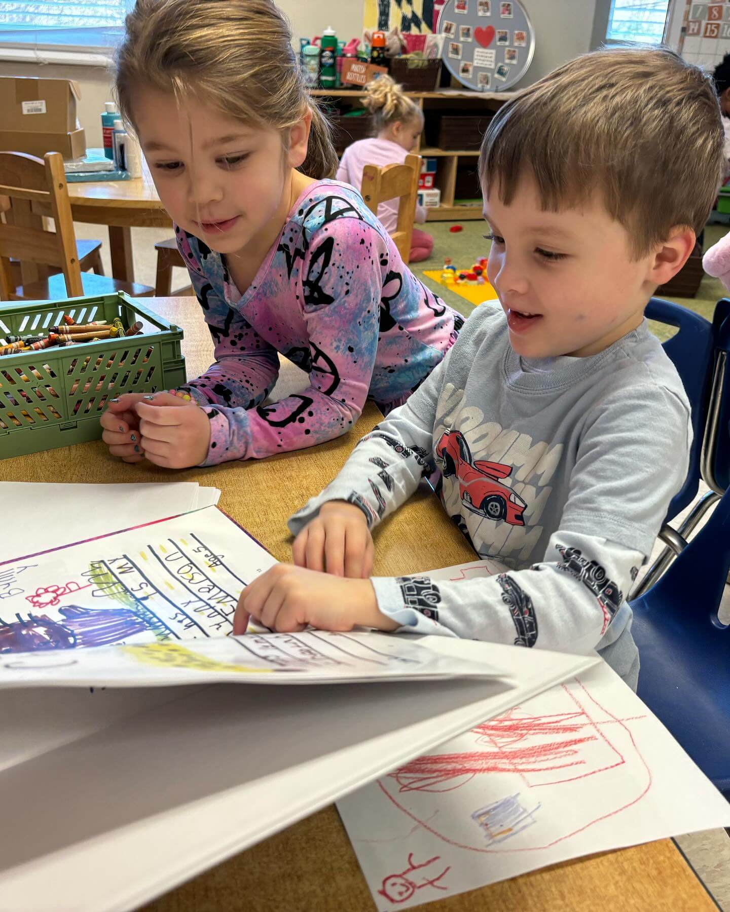 Kindergarten_2 Two children looking through a large workbook together, with crayons and drawings on the table.
