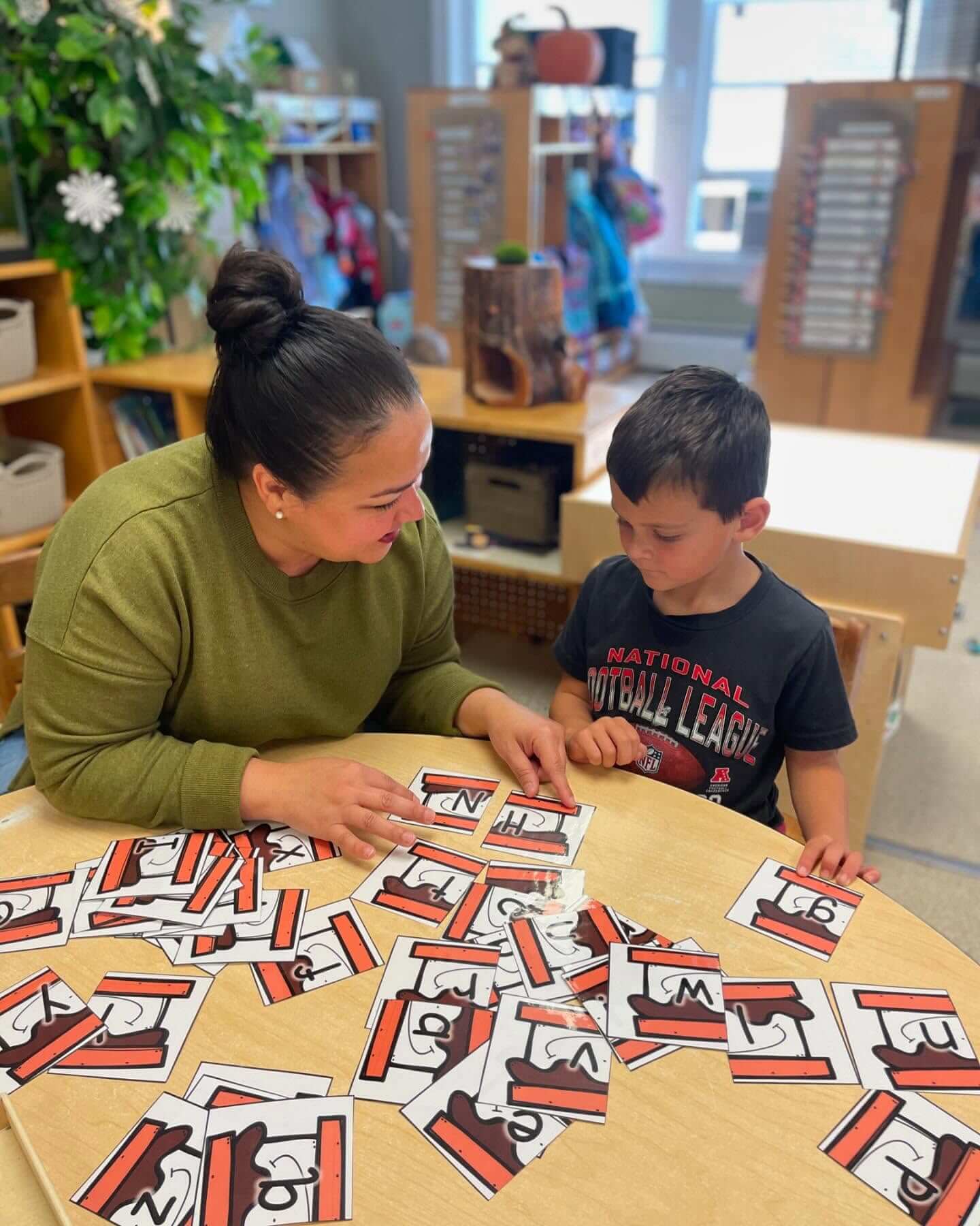 Kindergarten_4 Teacher working one-on-one with a young boy at a round table, matching number cards in a classroom setting.