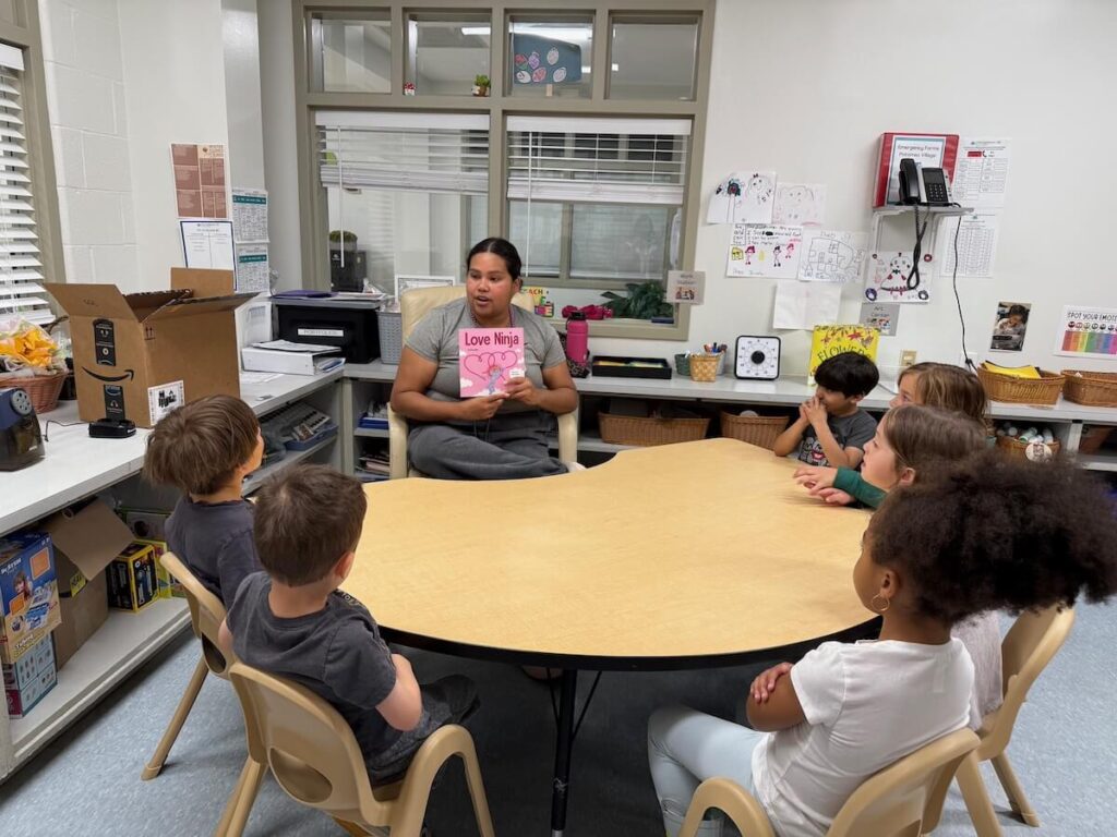 Teacher reading a book titled “Love Ninja” to a group of children seated around a table.