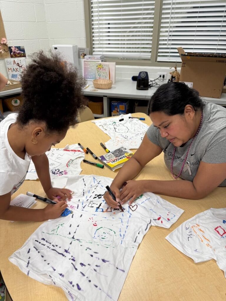 Teacher and child decorating a white T-shirt together with colorful marker drawings.