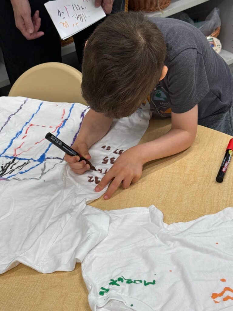 Child leaning over a table, drawing on a white T-shirt with markers.