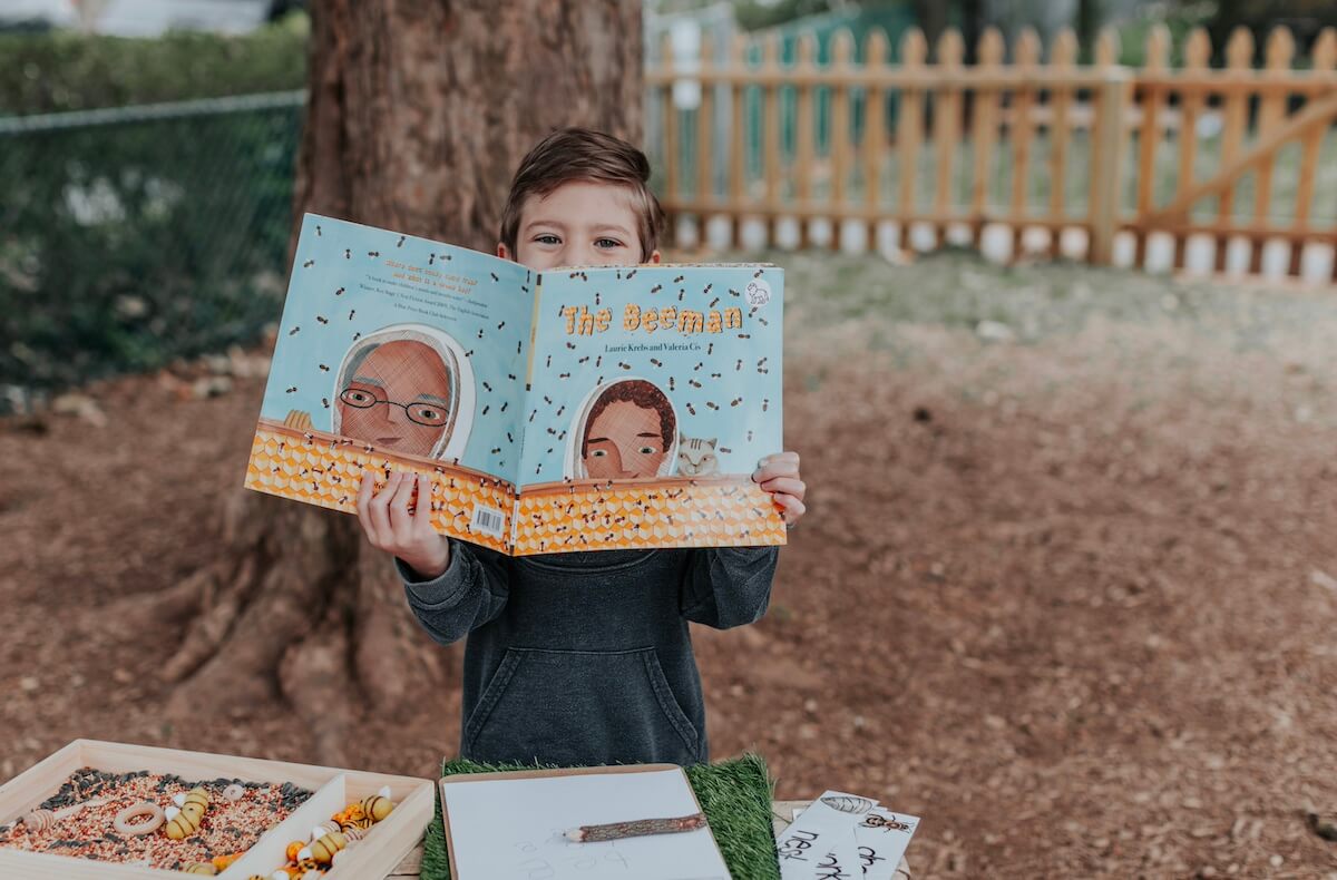 PreK_3 Child outdoors holding up a book titled ‘The Beeman’ while participating in a bee-themed learning activit