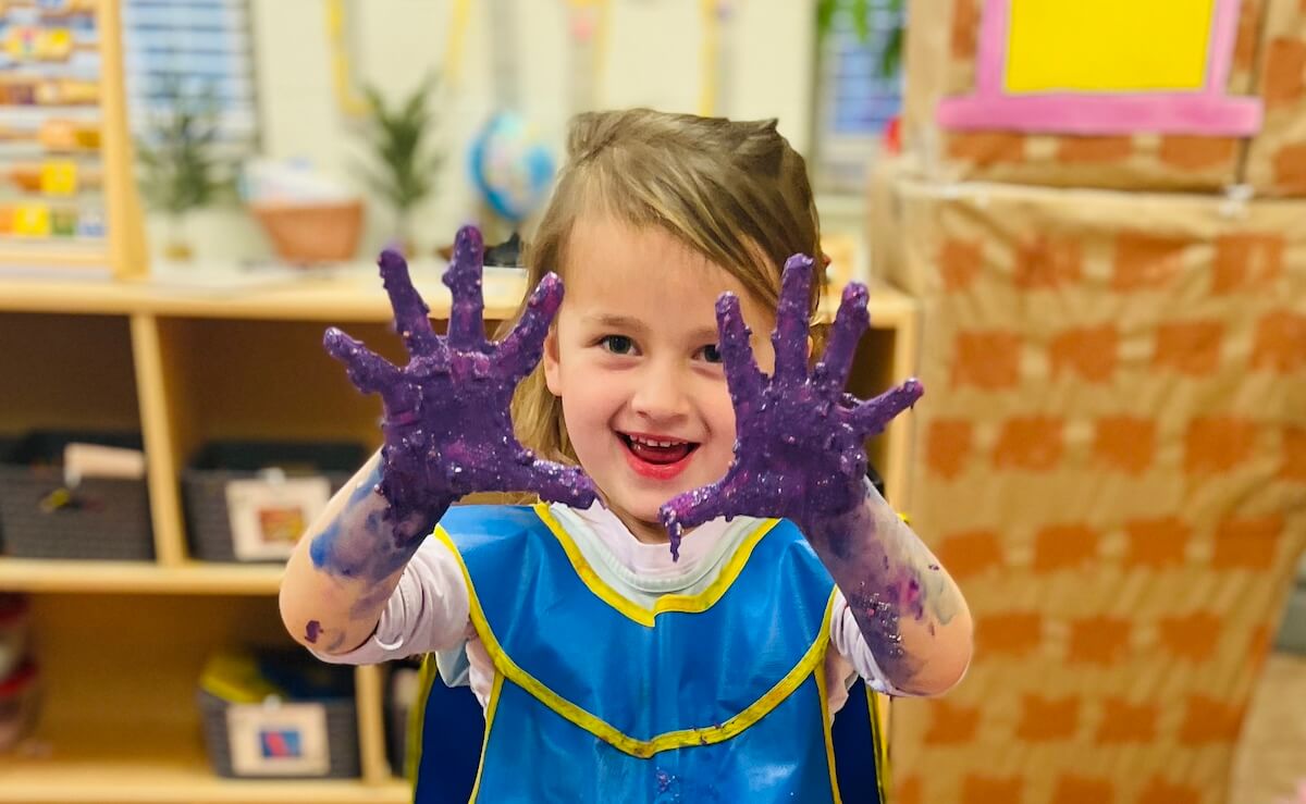 PreK_4 Preschool girl smiling with hands covered in purple paint during a messy art project.