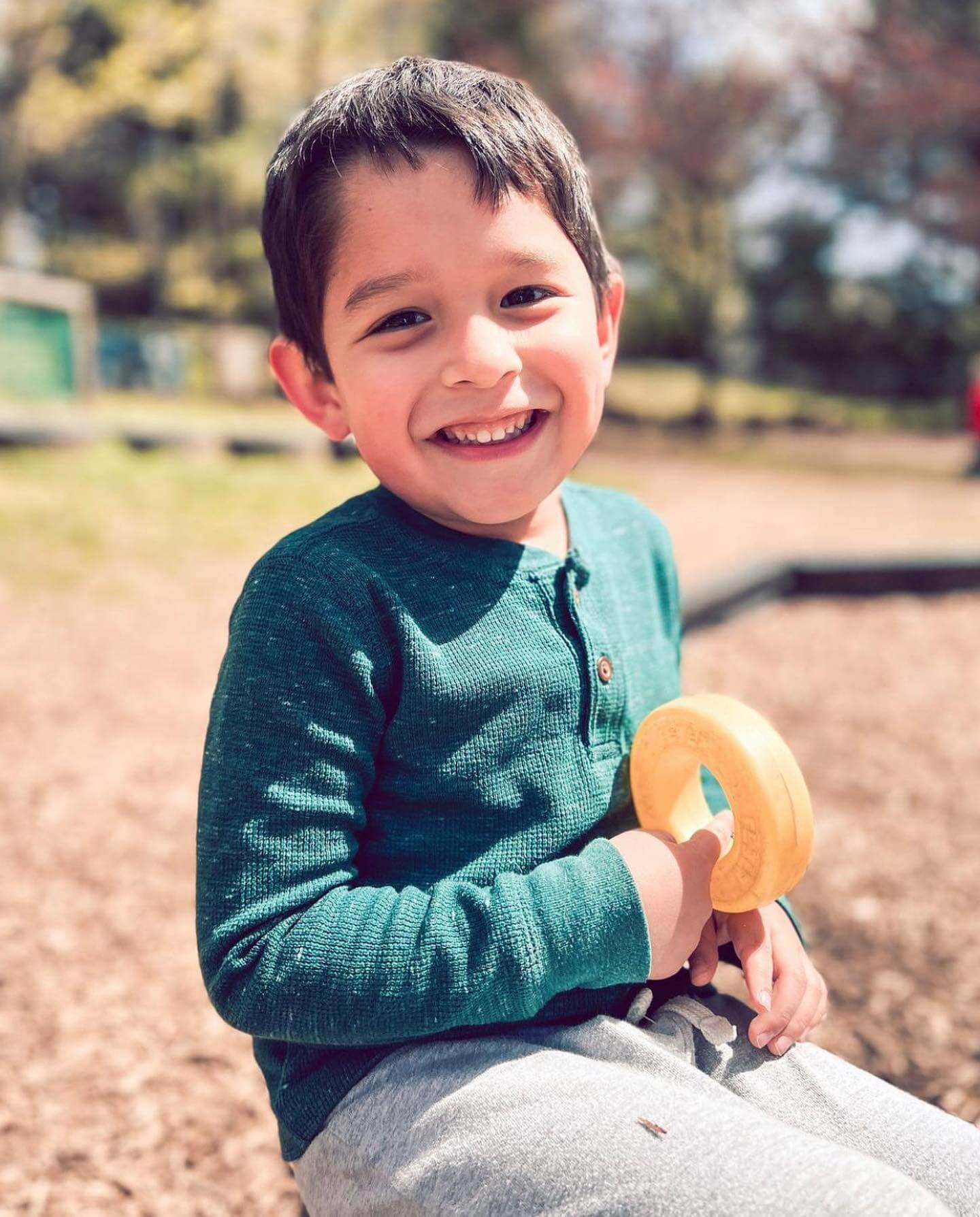 PreK_7 Preschool boy in a green shirt holding a yellow ring toy and smiling during outdoor playtime.