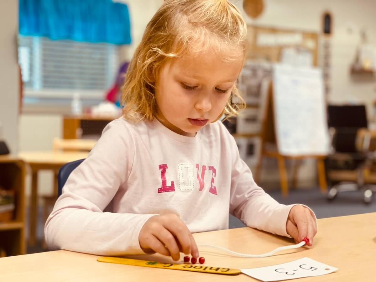 TK-6 Young girl in a classroom focusing on threading a string through a number card during a hands-on learning activity.