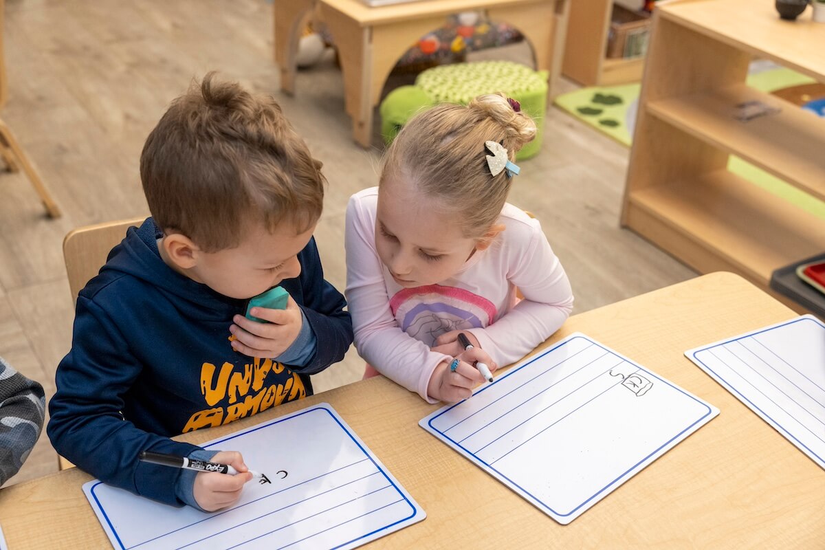 TK_1 Two young children sitting at a classroom table, working together on whiteboards with dry erase markers, practicing writing letters.