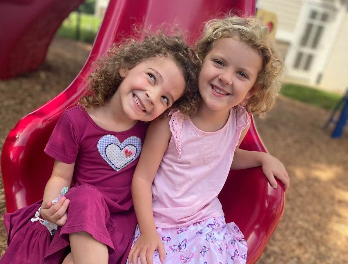 TK_3 Two smiling girls sitting together at the bottom of a bright red playground slide, one leaning her head on the other’s shoulder.