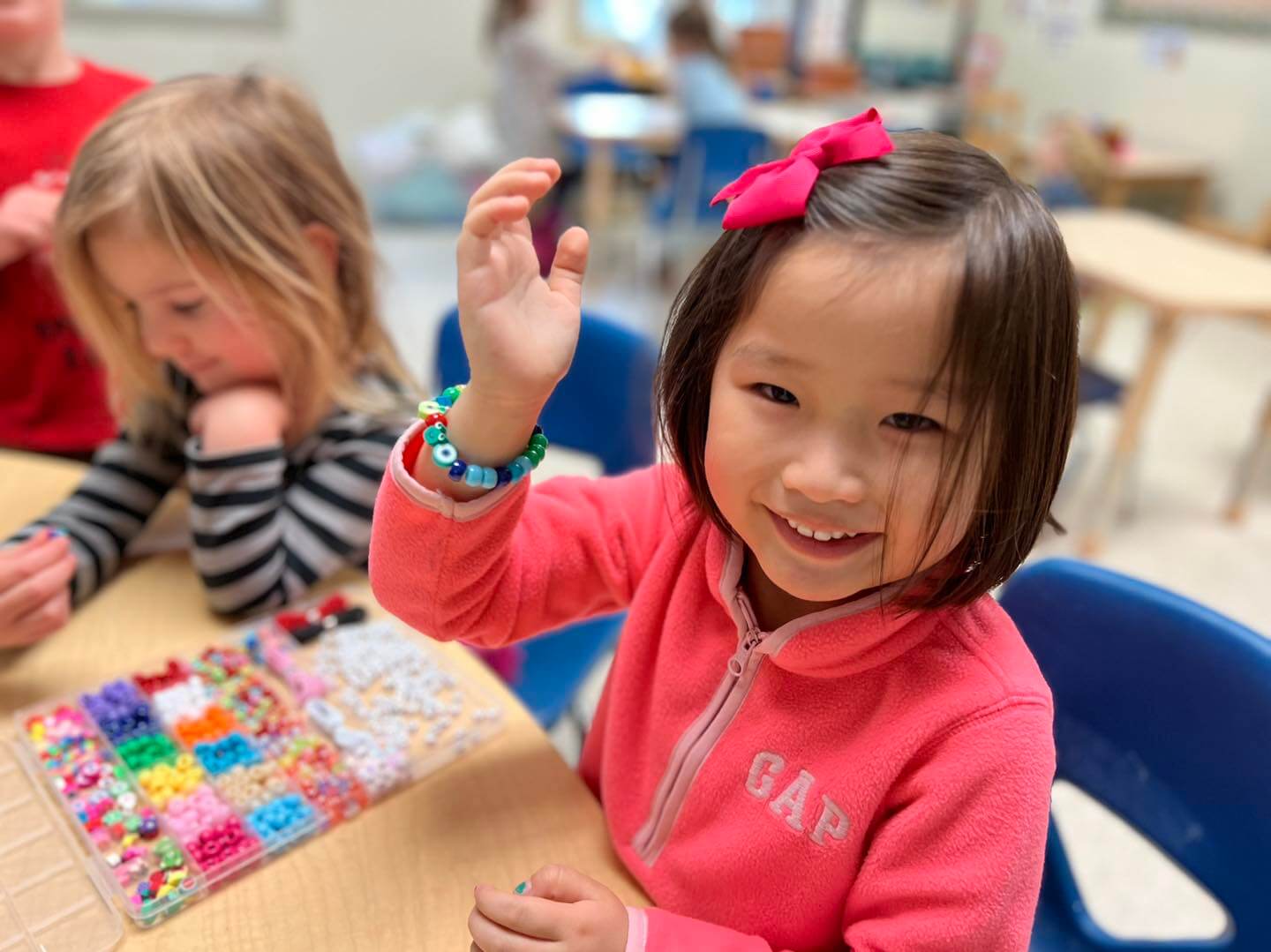 Threes_4 Girl in a pink fleece jacket with a bow in her hair showing a handmade beaded bracelet during a preschool art project.