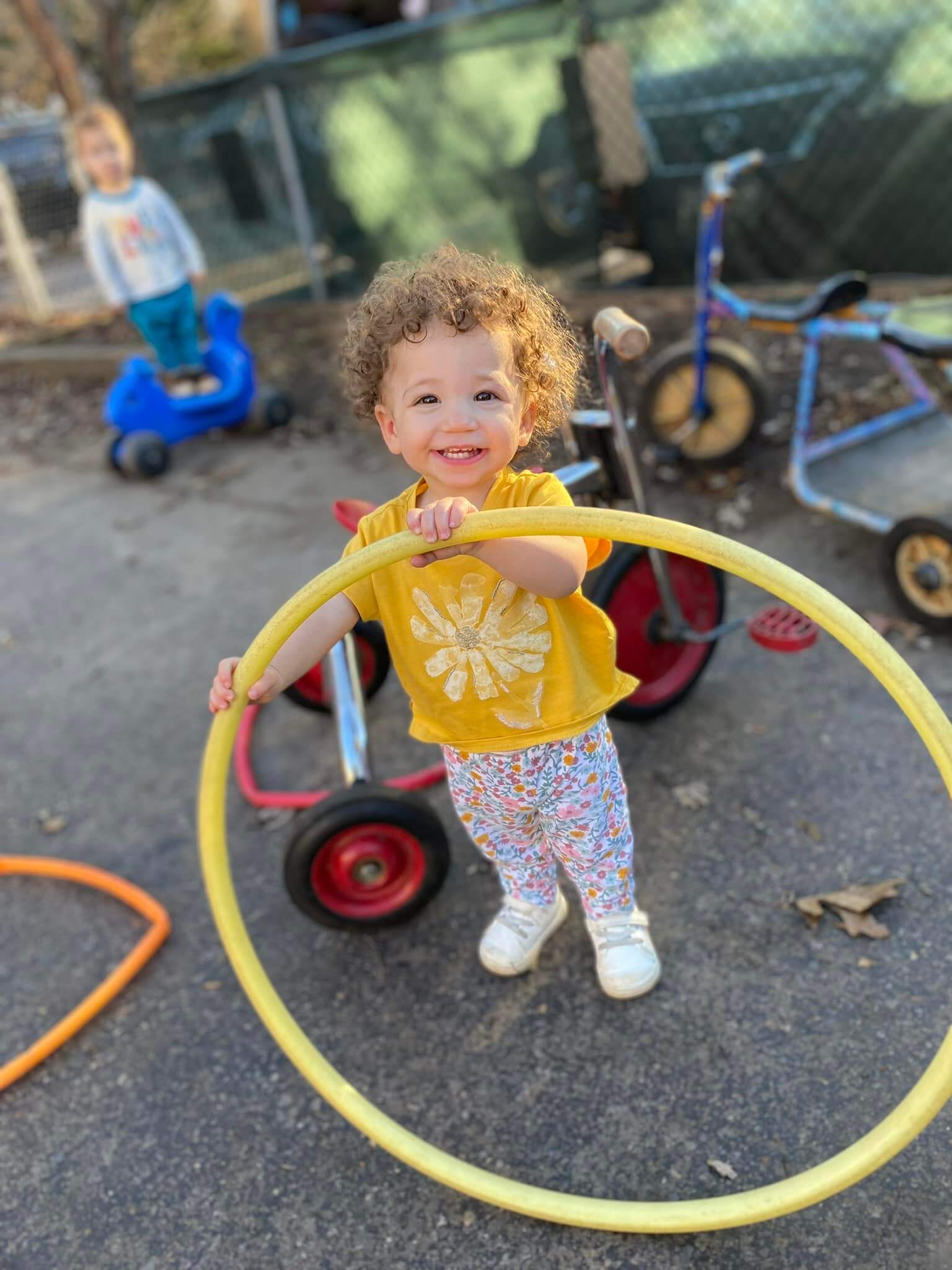 Twos_1 Smiling toddler in a yellow shirt holding a yellow hula hoop outside, with tricycles and another child in the background