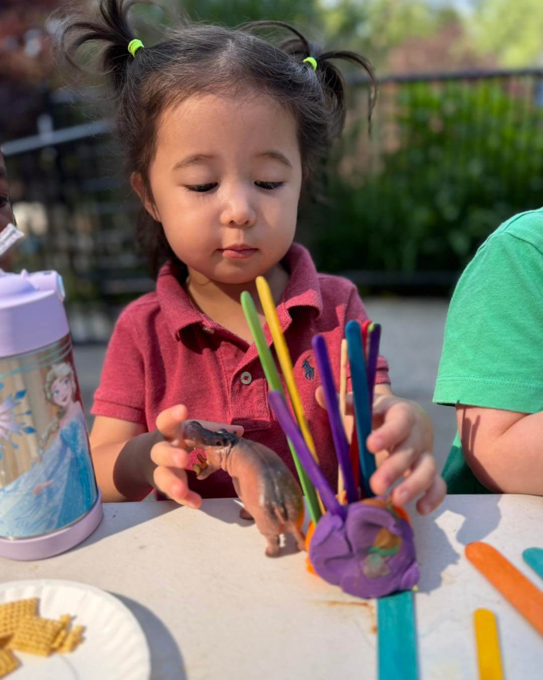 Twos_3 Child in a red shirt creating a craft with colorful popsicle sticks and playdough at an outdoor table.